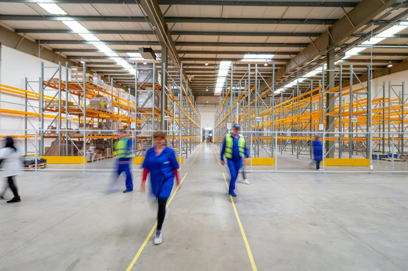 Workers walking through a warehouse aisle lined with stacked goods